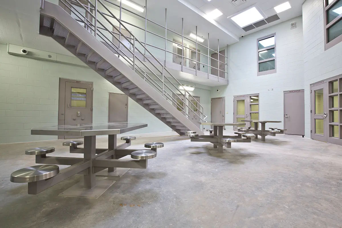 Spacious jail common area with metal tables and stools, surrounded by cell doors. A metal staircase leads to an upper level. Bright and utilitarian.