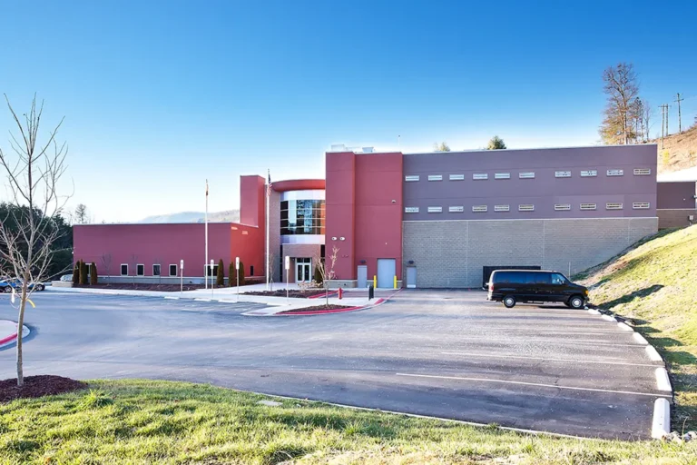 Modern building with red and beige walls, surrounded by a spacious parking lot on a sunny day. A black van is parked, and hills are visible in the background.