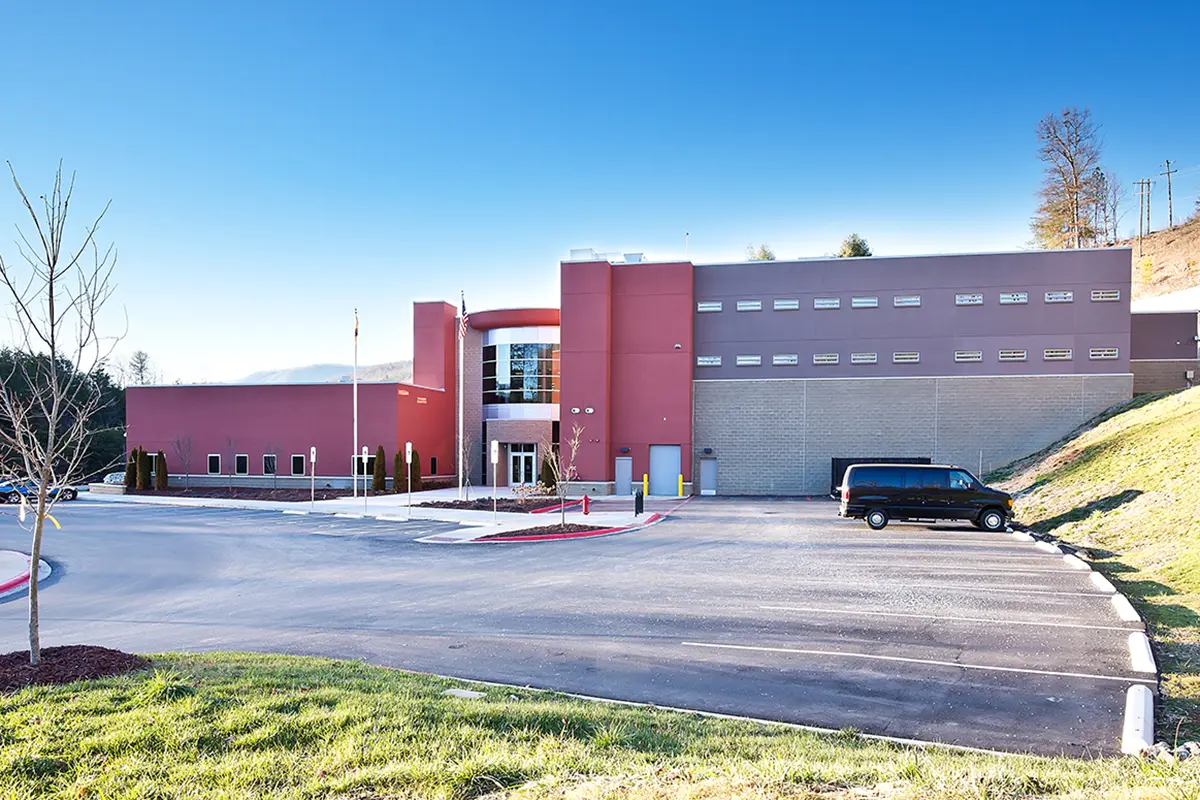 Modern building with red and beige walls, surrounded by a spacious parking lot on a sunny day. A black van is parked, and hills are visible in the background.