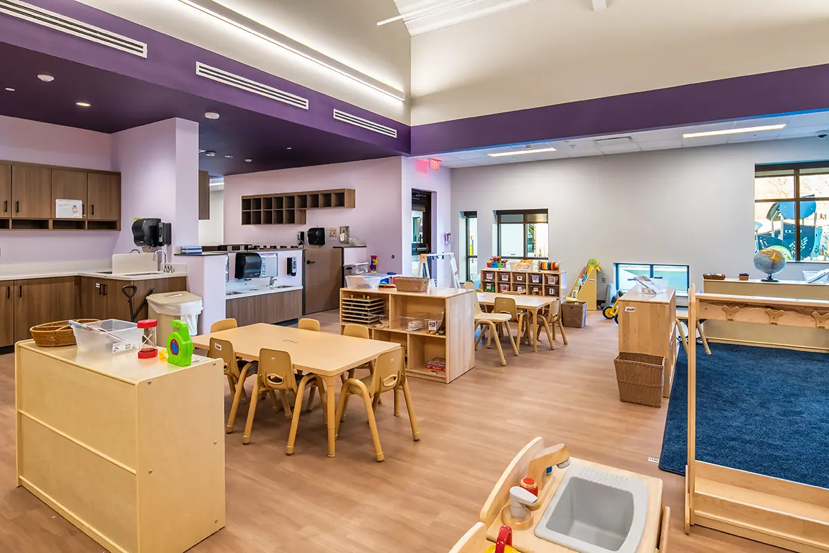 A bright, colorful preschool classroom with wood tables, chairs, and storage. Playspaces, a toy kitchen, and a cozy rug create a welcoming atmosphere.