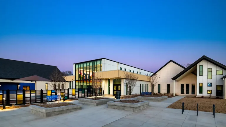 Modern building at dusk with geometric architecture, large glass windows, and an inviting courtyard. The sky is a gradient of blue and purple, creating a serene atmosphere.