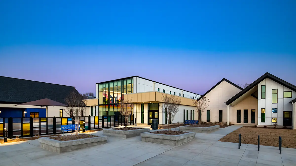 Modern building at dusk with geometric architecture, large glass windows, and an inviting courtyard. The sky is a gradient of blue and purple, creating a serene atmosphere.