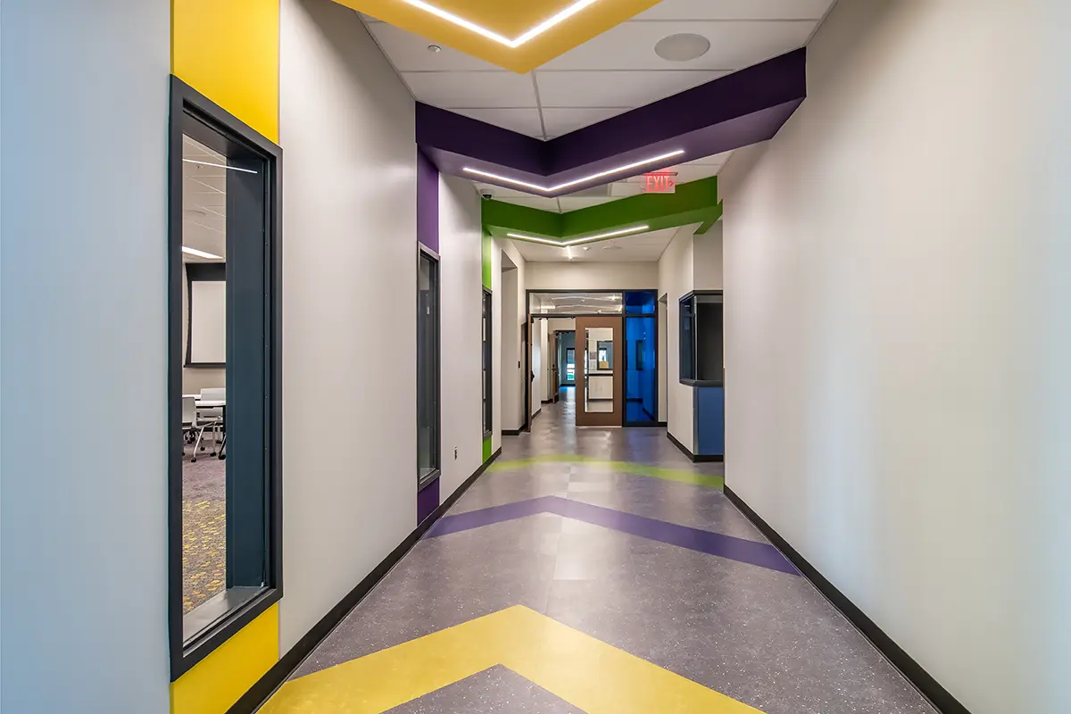 A hallway with vibrant colors features purple, yellow, and green geometric accents on walls and ceiling. Large windows line the left, creating a modern, lively atmosphere.