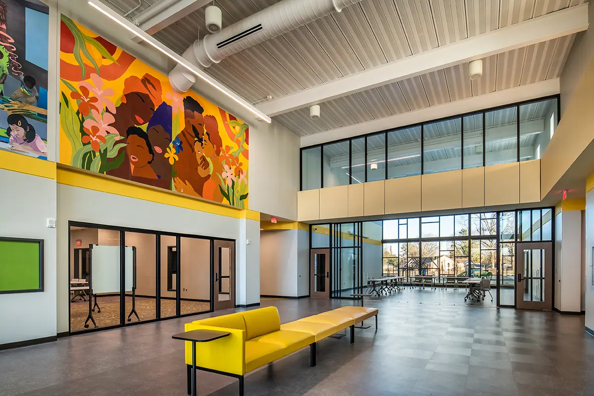 Spacious, modern lobby with high ceilings and large windows, vibrant mural depicting diverse faces and floral designs. Bright atmosphere, yellow benches.