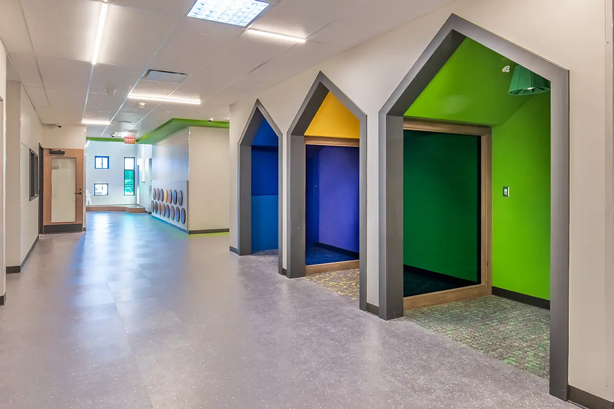 A modern hallway with gray flooring and bright, colorful cubbies. Each cubby has a pointed arch design, painted in green, blue, and yellow tones. The space feels vibrant and welcoming.