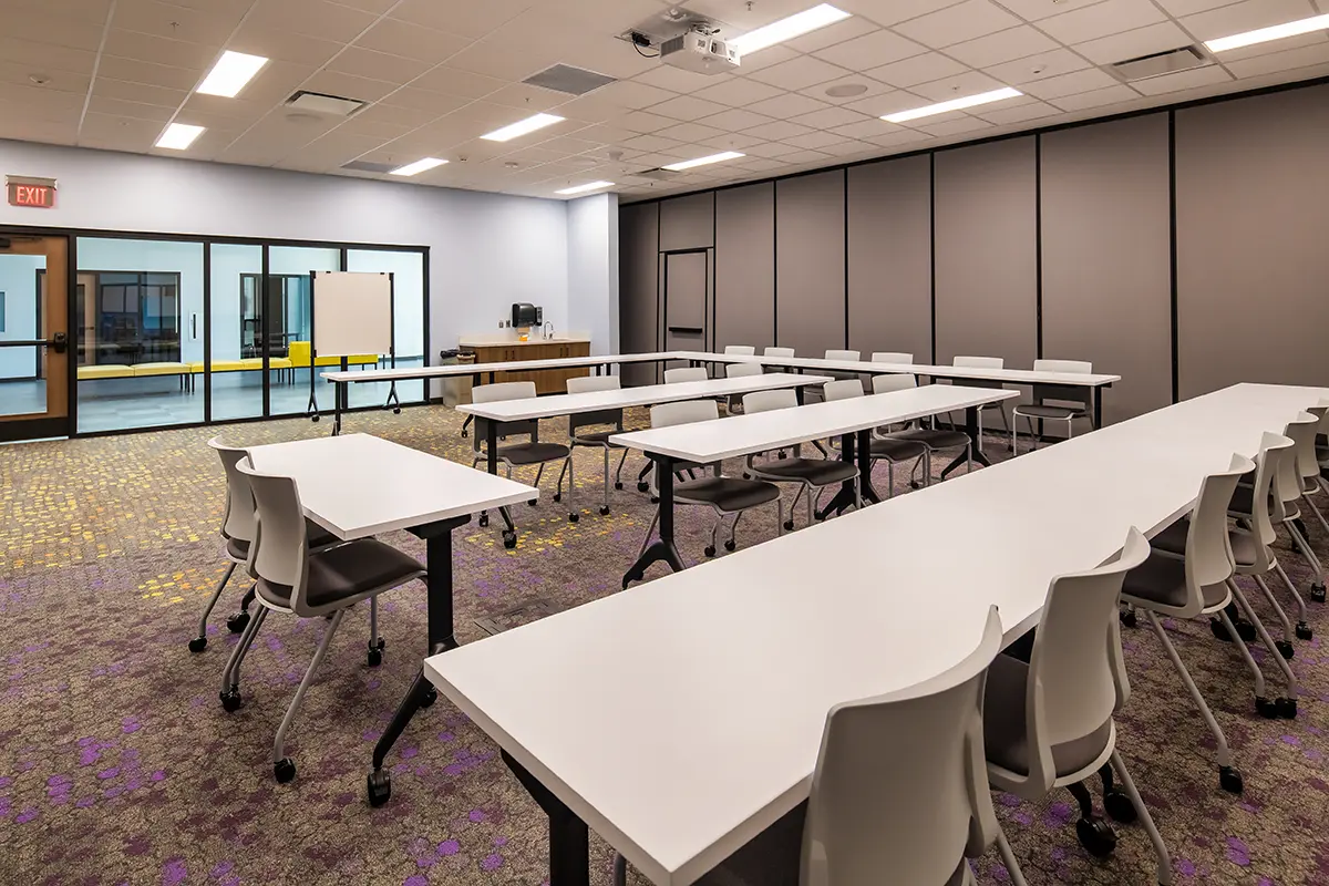 Modern conference room with white tables in U-shape, gray chairs, projector on ceiling, and sliding glass doors. The atmosphere is professional and organized.