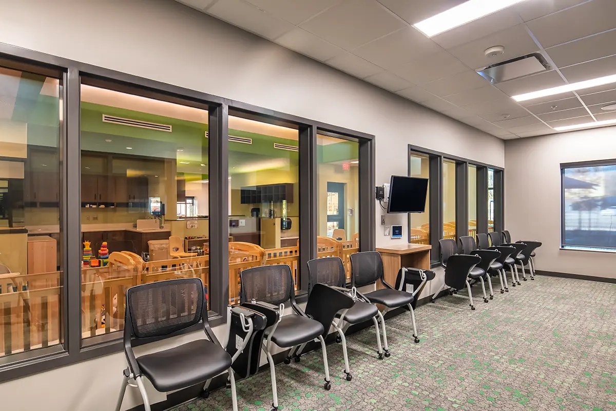 A daycare observation room with chairs along a wall, facing large windows that reveal a playroom with colorful toys and cribs, conveying a serene setting.