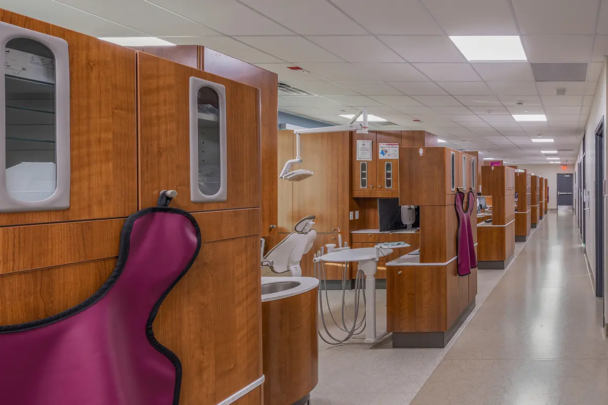 Long corridor of a dental clinic with multiple wooden partitioned stations. Each station has dental equipment and pink protective aprons hang nearby. Bright, clean, clinical ambiance.