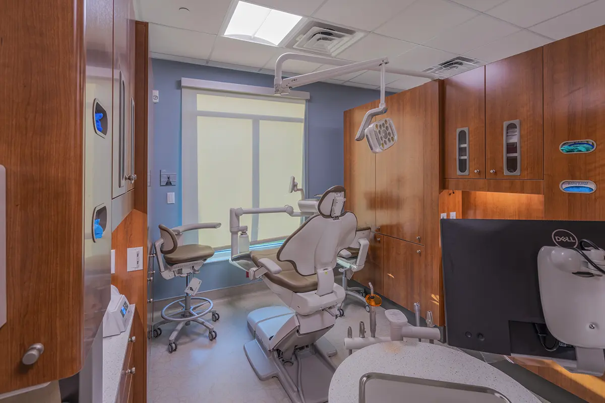 A modern dental office with a reclining dental chair, overhead light, and wooden cabinets. The room is brightly lit, creating a clean and professional atmosphere.