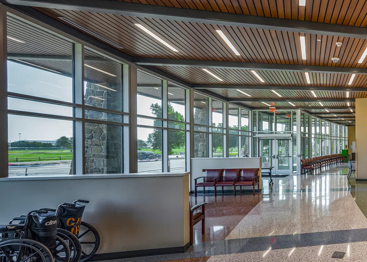 Modern lobby with large floor-to-ceiling windows, showing greenery outside. The space has a wood-paneled ceiling, glossy floor, and red seating. Two wheelchairs are in the foreground. Bright and welcoming atmosphere.