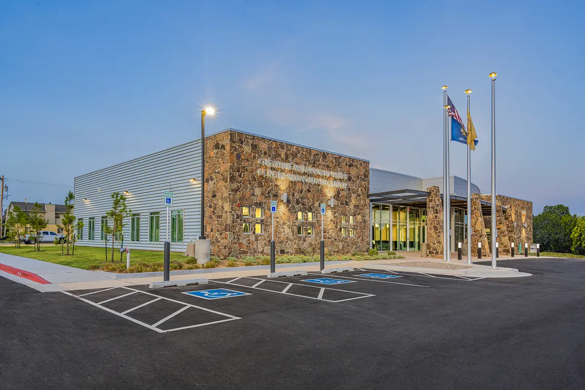 Modern public building at dusk with stone facade and large windows, surrounded by parking spaces and flagpoles, conveying a sense of community.