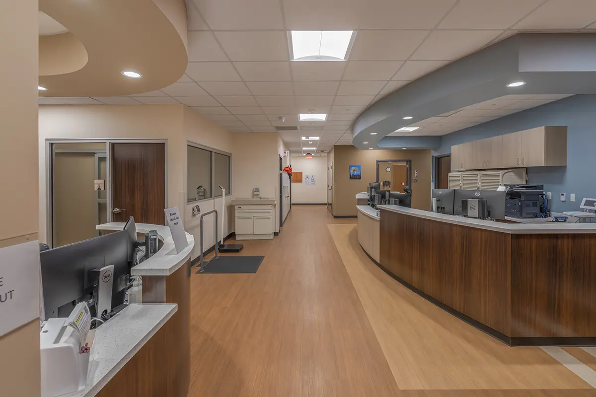 Modern hospital reception area with clean lines and neutral tones. The curving wooden desk has computers; the hallway extends into the background, conveying a calm, organized atmosphere.