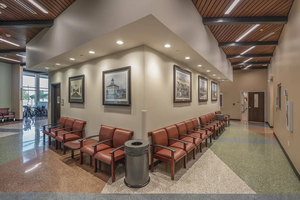 A spacious waiting area with rows of red cushioned chairs lining two walls. Art prints are displayed on cream walls, and the ceiling features wooden panels.