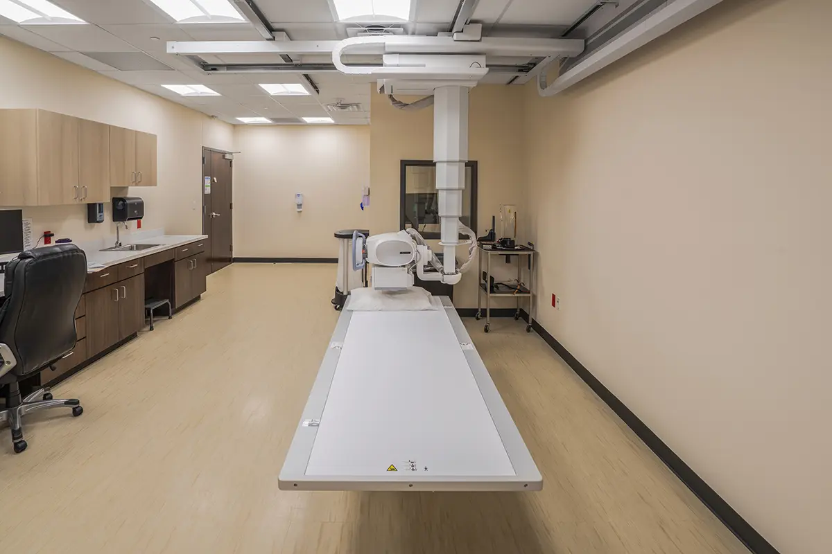 A clean, modern X-ray room with beige walls and vinyl flooring. An empty X-ray table is centered, with cabinets and a swivel chair on the left. The atmosphere is clinical.
