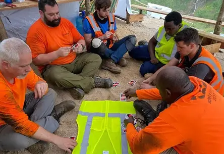 A group of six construction workers sits on sandy ground, engaged in a lively card game. They are wearing bright orange and blue vests, creating a friendly and relaxed atmosphere.