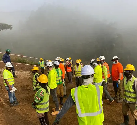 A group of construction workers in safety gear and helmets stand on a foggy hillside. They appear to be engaged in a discussion, creating a focused atmosphere.