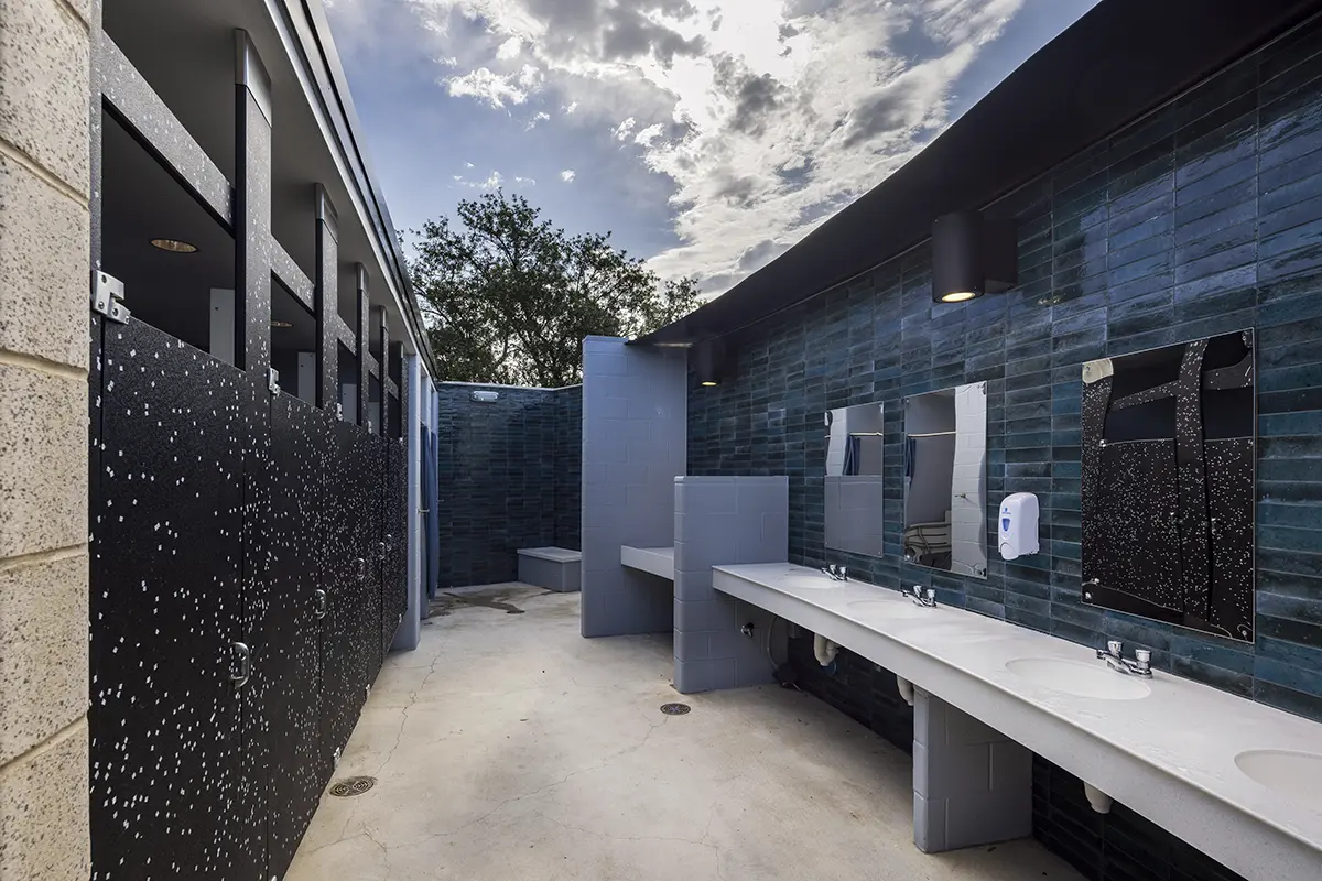 Modern restroom with sleek blue-tiled walls, concrete sinks, and black speckled stalls. Open ceiling under cloudy sky, creating an airy atmosphere.