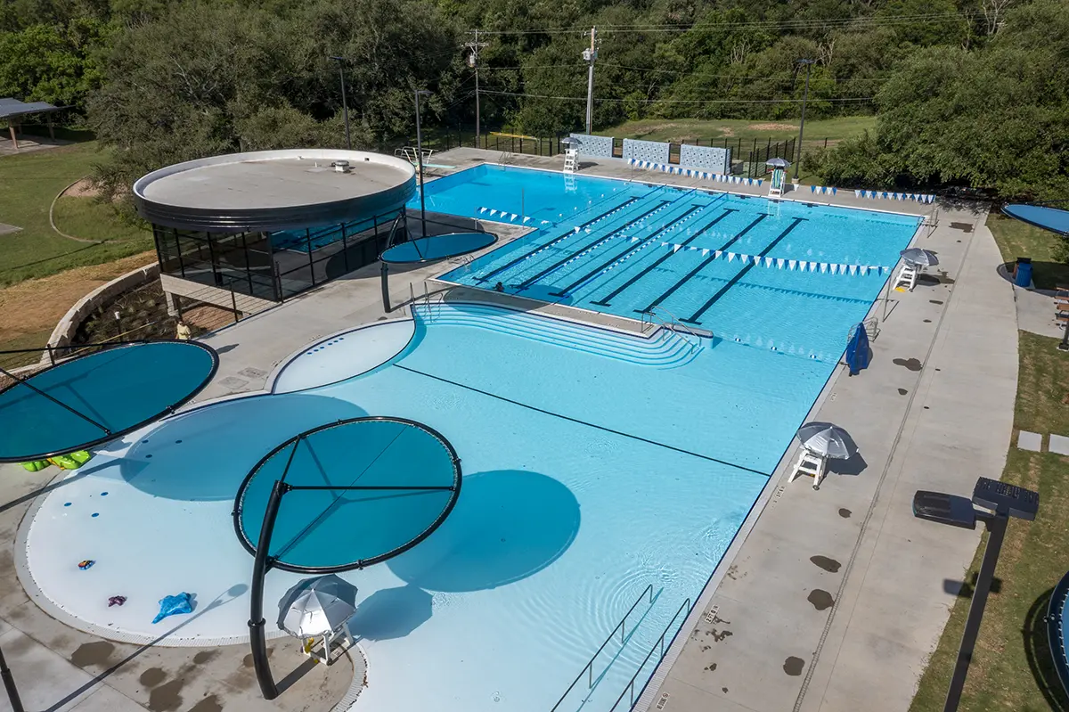 Aerial view of an outdoor swimming pool with lap lanes, round shaded areas, and a circular building. Surrounded by greenery, creating a serene ambiance.
