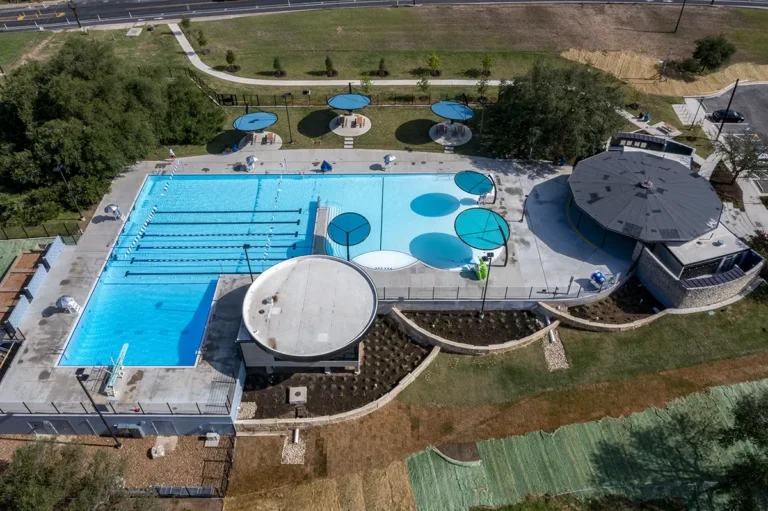 Aerial view of a public swimming pool complex with lanes, umbrellas for shade, a grassy area, and surrounding pathways, conveying a recreational vibe.