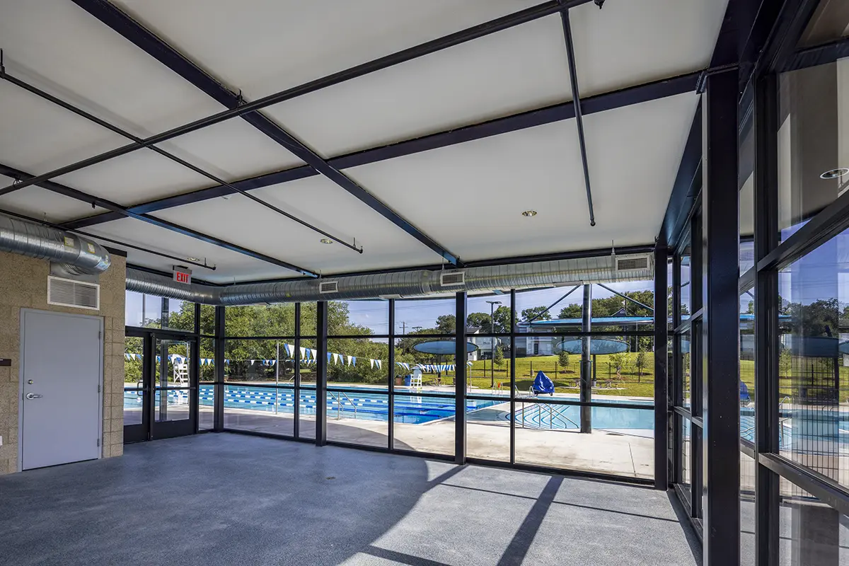 Indoor pool view from a glass-walled room, showcasing an empty swimming pool with blue checkered flags outside. Bright, sunny atmosphere.
