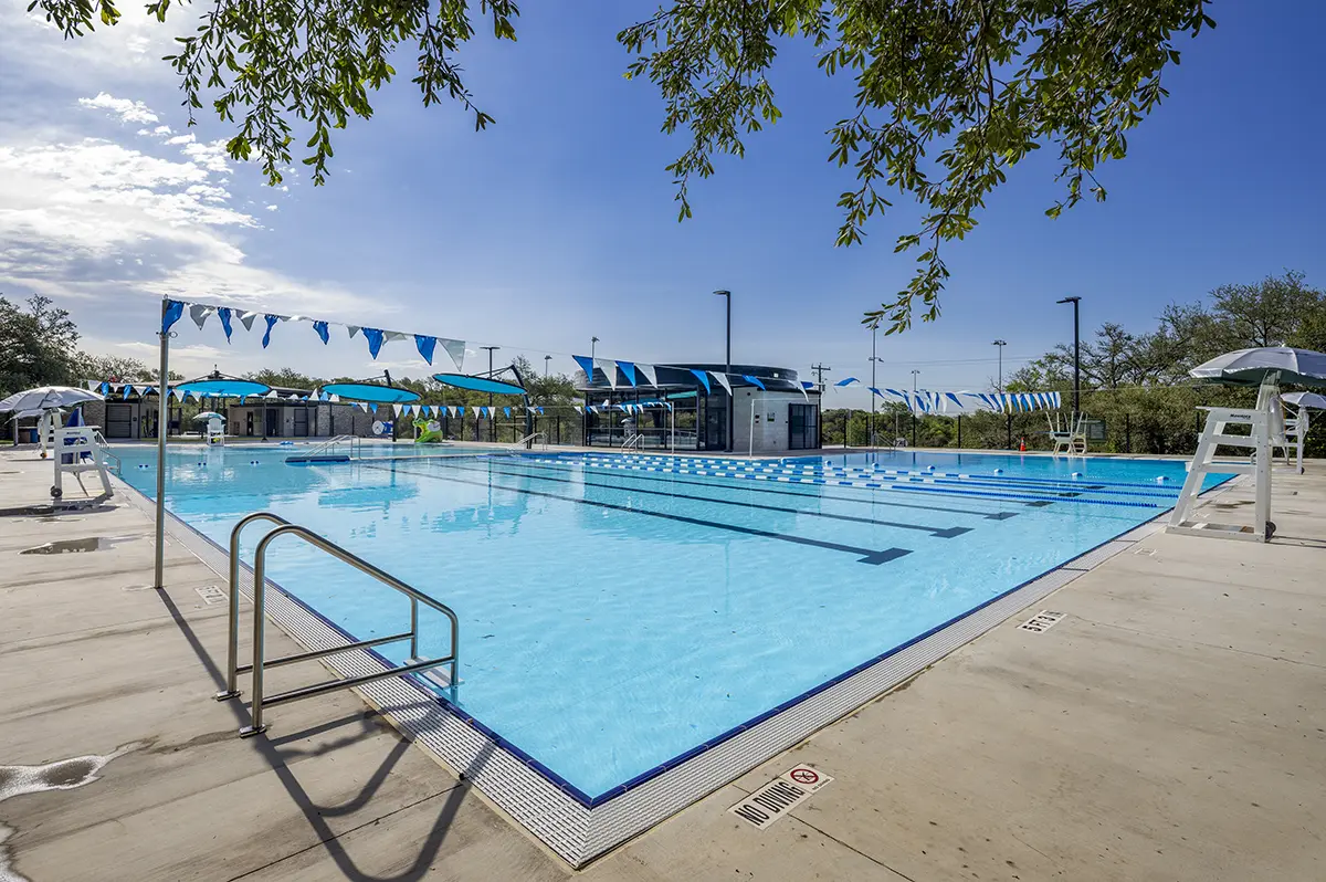 Outdoor swimming pool with clear blue water, surrounded by sunlit deck, lifeguard chairs, and shaded bleachers. Blue sky and leafy trees set a tranquil scene.