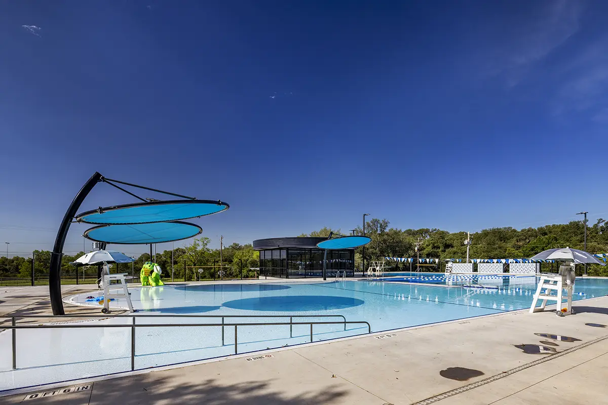Outdoor swimming pool with blue shade structures and lifeguard chairs, under a clear blue sky. Greenery in the background creates a serene setting.