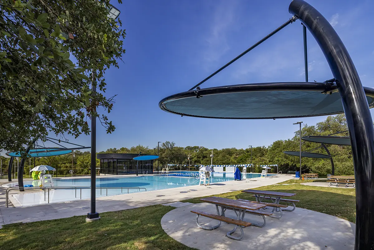 Outdoor pool area under a clear blue sky, with modern circular shades, picnic tables on grass, and a serene atmosphere inviting relaxation.