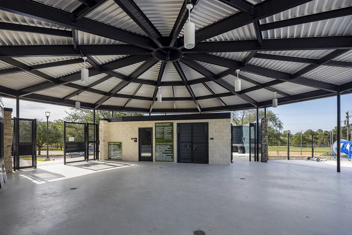 A spacious pavilion with a circular metal roof and hanging lights. Open sides reveal trees, fences, and a sports field, conveying an airy, communal feel.
