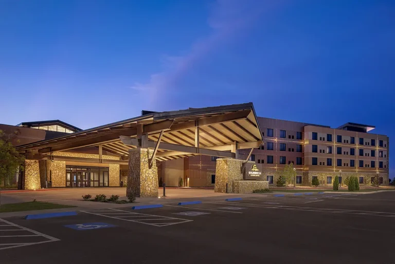 Lit building entrance at dusk, featuring stone columns and a modern facade with a warm, inviting glow against a deep blue sky. Quiet parking area in front.