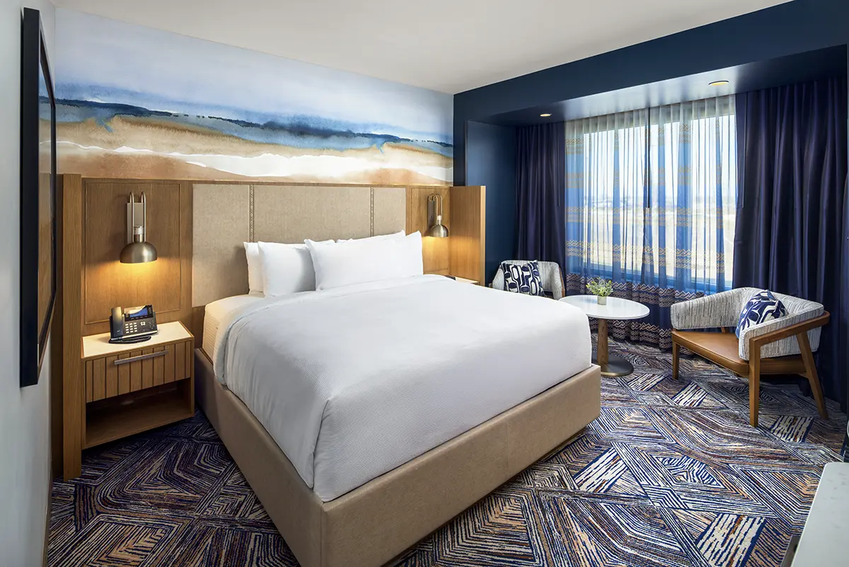 Modern hotel suite with a green and wood theme. Features a cozy seating area, a desk with a TV, and a bed framed by a large scenic artwork.