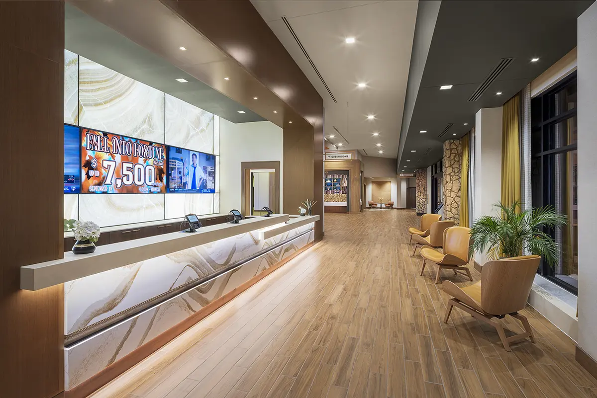 Modern hotel lobby with wooden floors, warm lighting, and a long reception desk. A digital screen advertises a $7,500 offer. Seating area with plants.