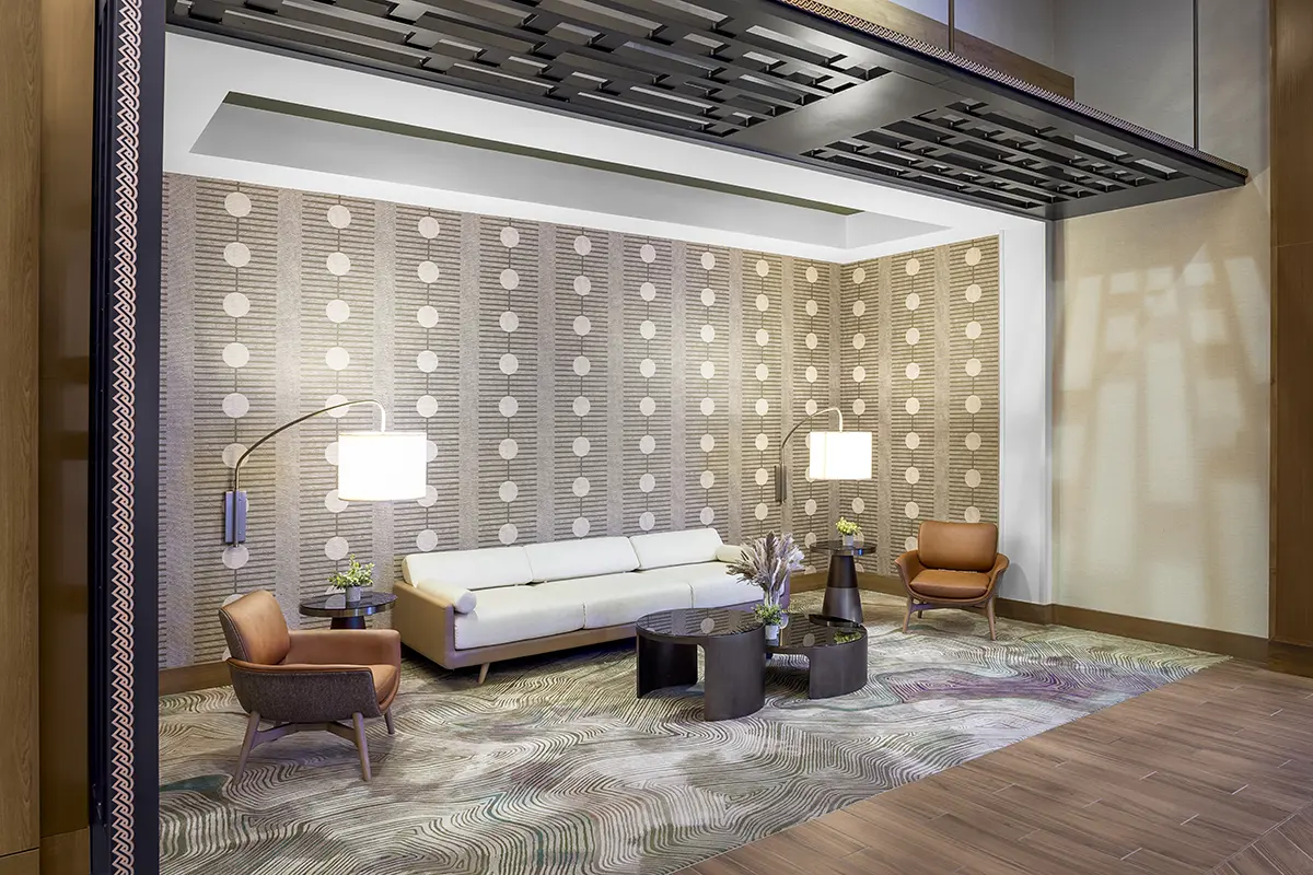 Modern hotel lobby with patterned wallpaper, beige couch, two brown chairs, and round tables. Soft lighting creates a warm, inviting atmosphere.