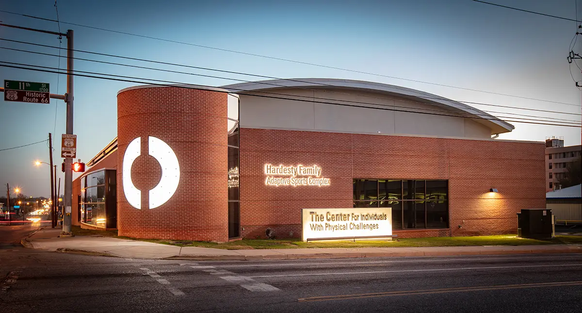 A modern brick building with a curved facade at dusk, featuring "Hardesty Family Adaptive Sports Complex" signage and glowing streetlights, creating a welcoming atmosphere.