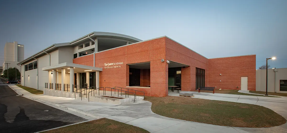 Modern brick and concrete building at dusk with large windows and an accessible ramp. The setting is serene, with soft exterior lighting and a clear sky.