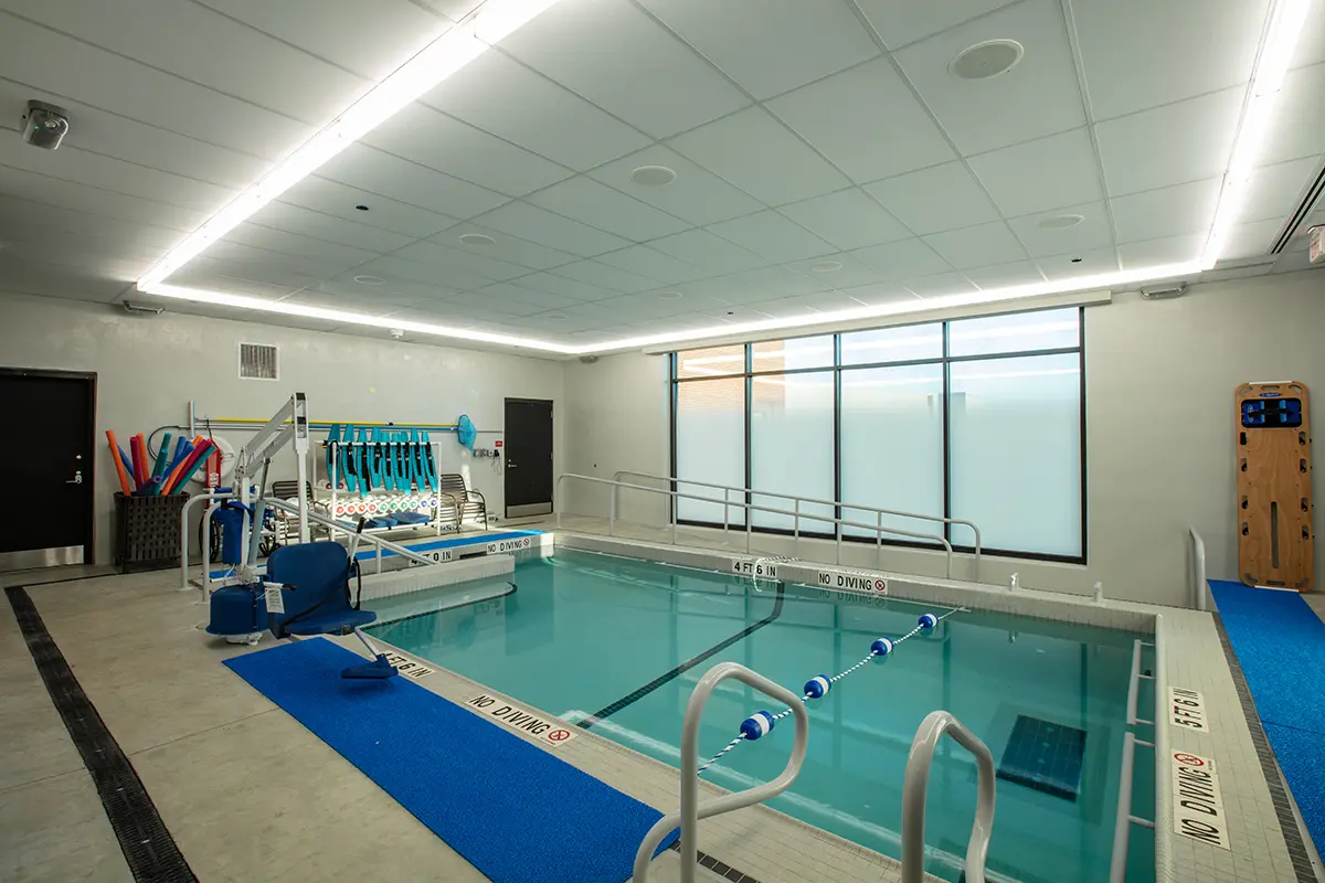 Indoor therapy pool with handrails, aquatic equipment, and no diving signs. Bright, modern setting emphasizes accessibility and relaxation.