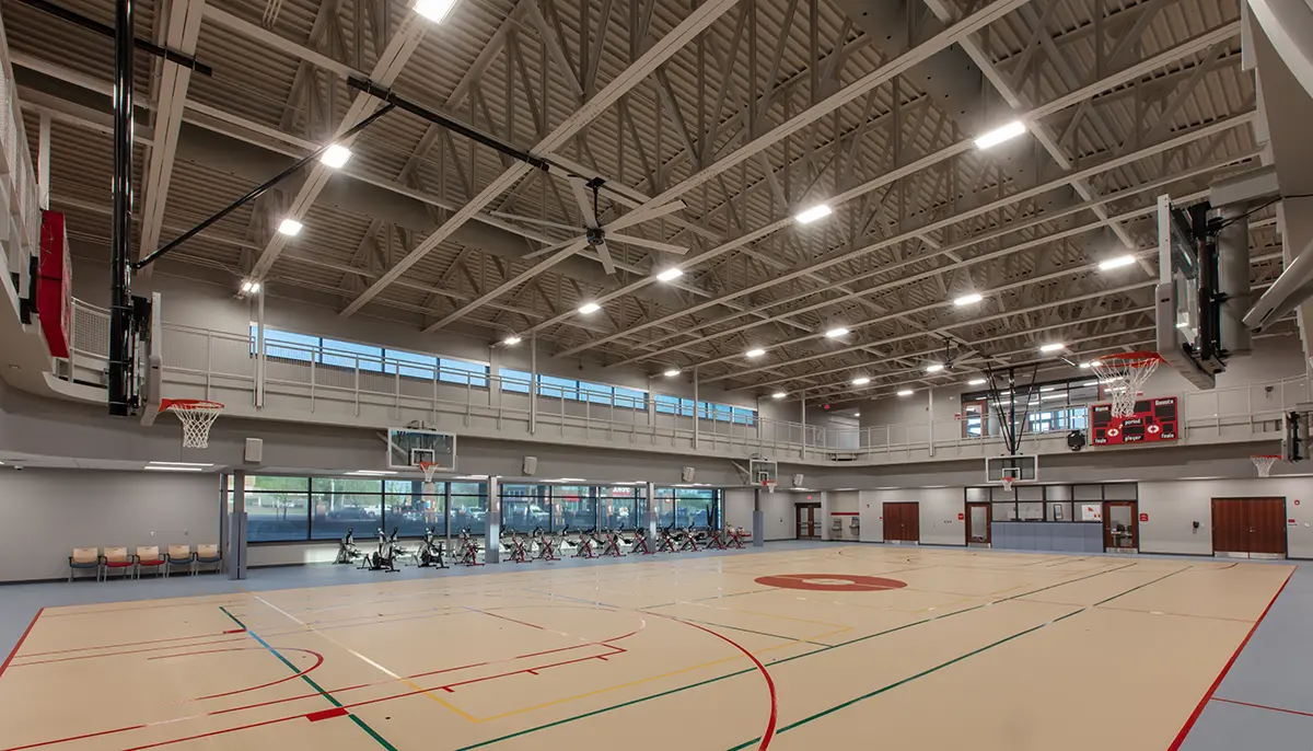 A spacious, well-lit gymnasium with high ceilings, multiple basketball hoops, and exercise bikes lined up along large windows. The atmosphere is bright and modern.