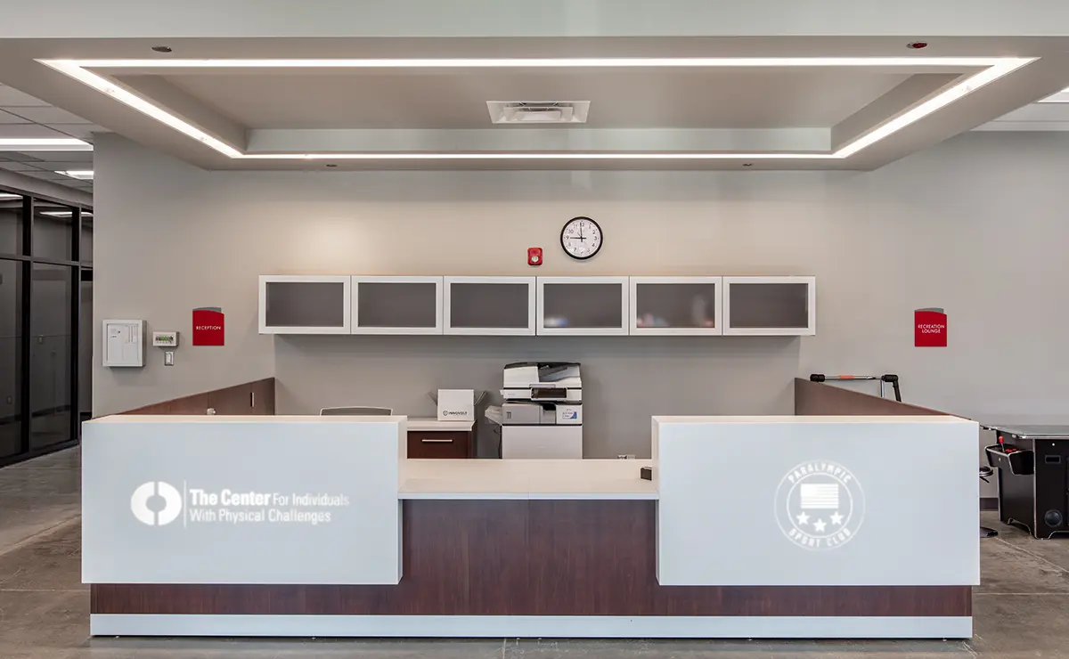 Modern reception area with a sleek desk, overhead cabinets, a clock, and office equipment. Logos on the desk convey an inclusive, professional tone.
