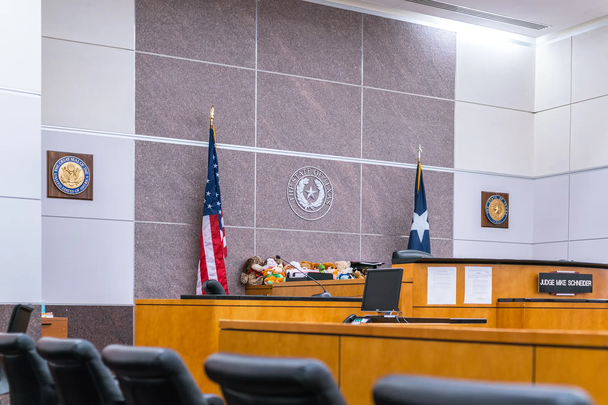 Sala de audiencias moderna con decoración en madera, asientos vacíos, estrado del juez con bandera y ordenadores. Iluminación brillante y ambiente formal y organizado.