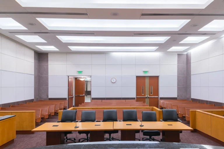 Empty courtroom with modern design, featuring wood-paneled walls, empty benches, a central table with chairs and microphones, and a wall clock.