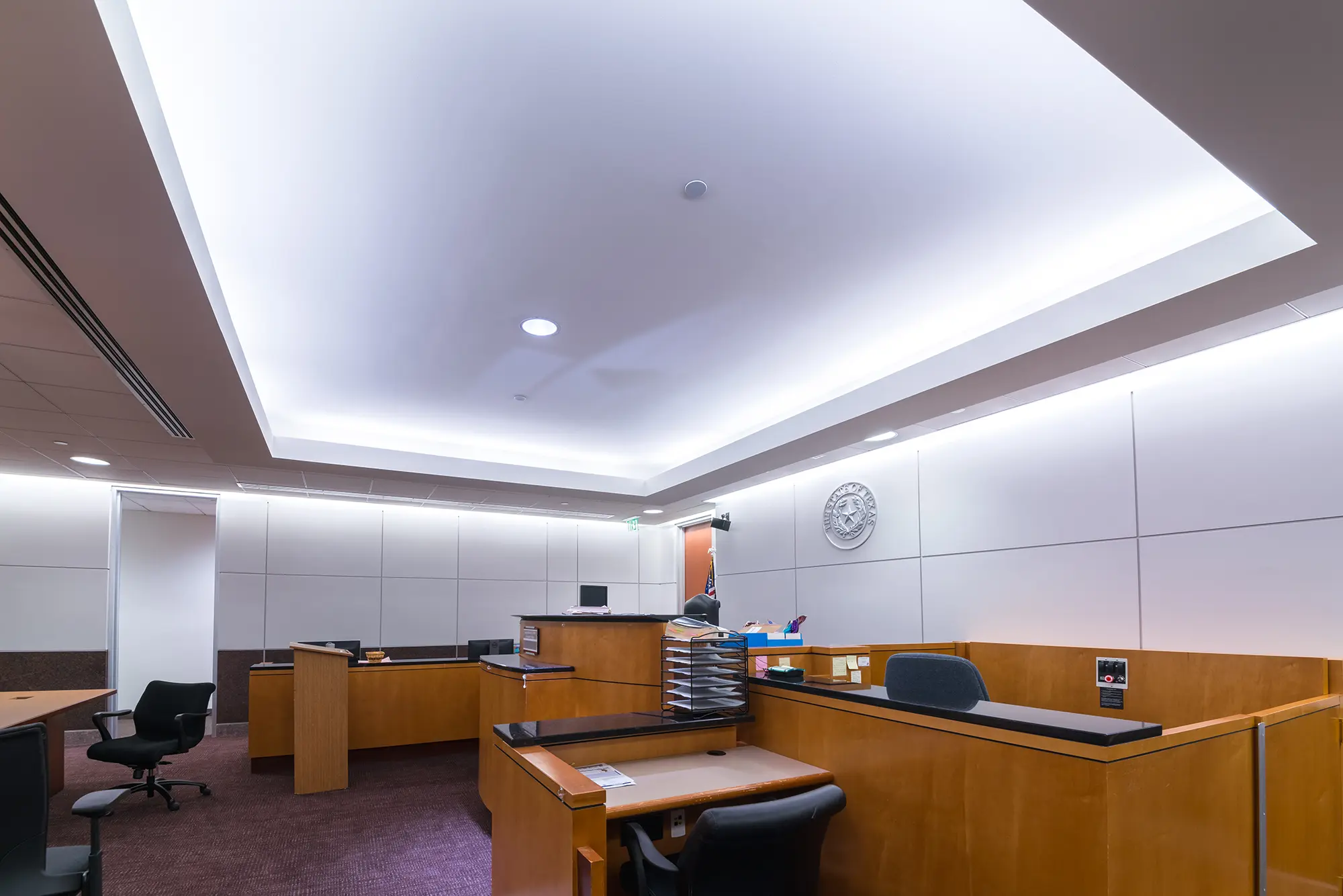 Sala de audiencias moderna con muebles de madera, sillas y un monitor de ordenador. Las luminosas luces del techo crean un ambiente formal y profesional.