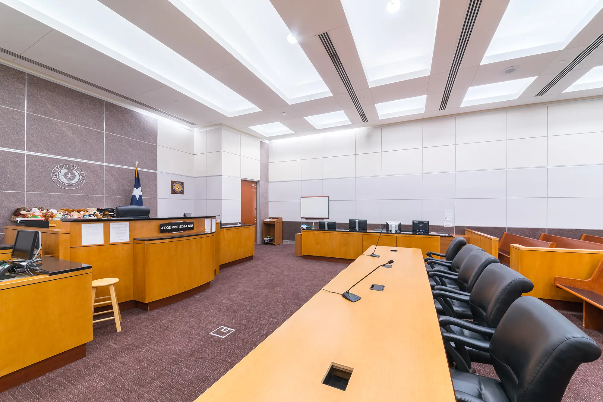 Sala de audiencias moderna con decoración en madera, asientos vacíos, estrado del juez con bandera y ordenadores. Iluminación brillante y ambiente formal y organizado.
