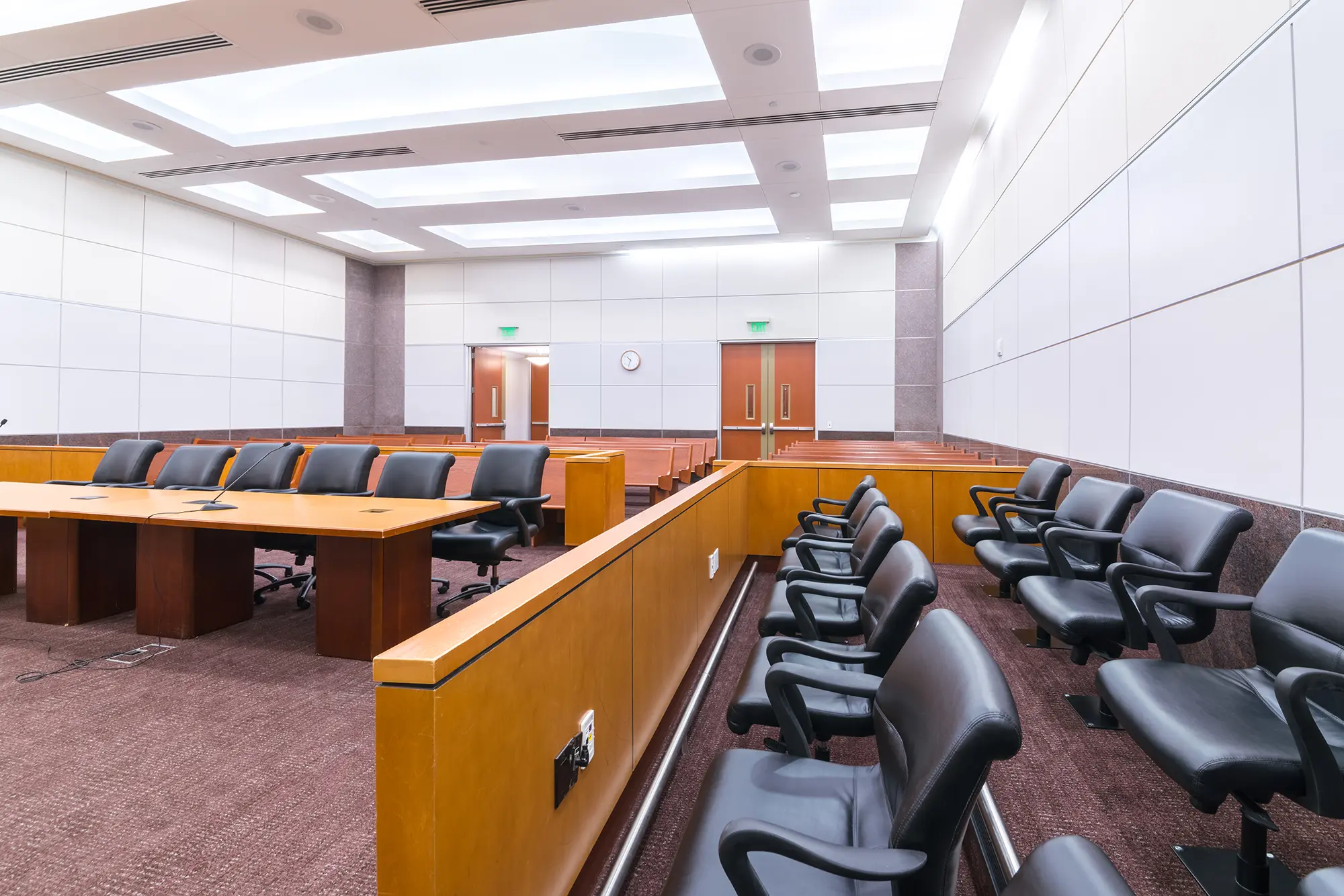 Sala de audiencias moderna con decoración en madera, asientos vacíos, estrado del juez con bandera y ordenadores. Iluminación brillante y ambiente formal y organizado.