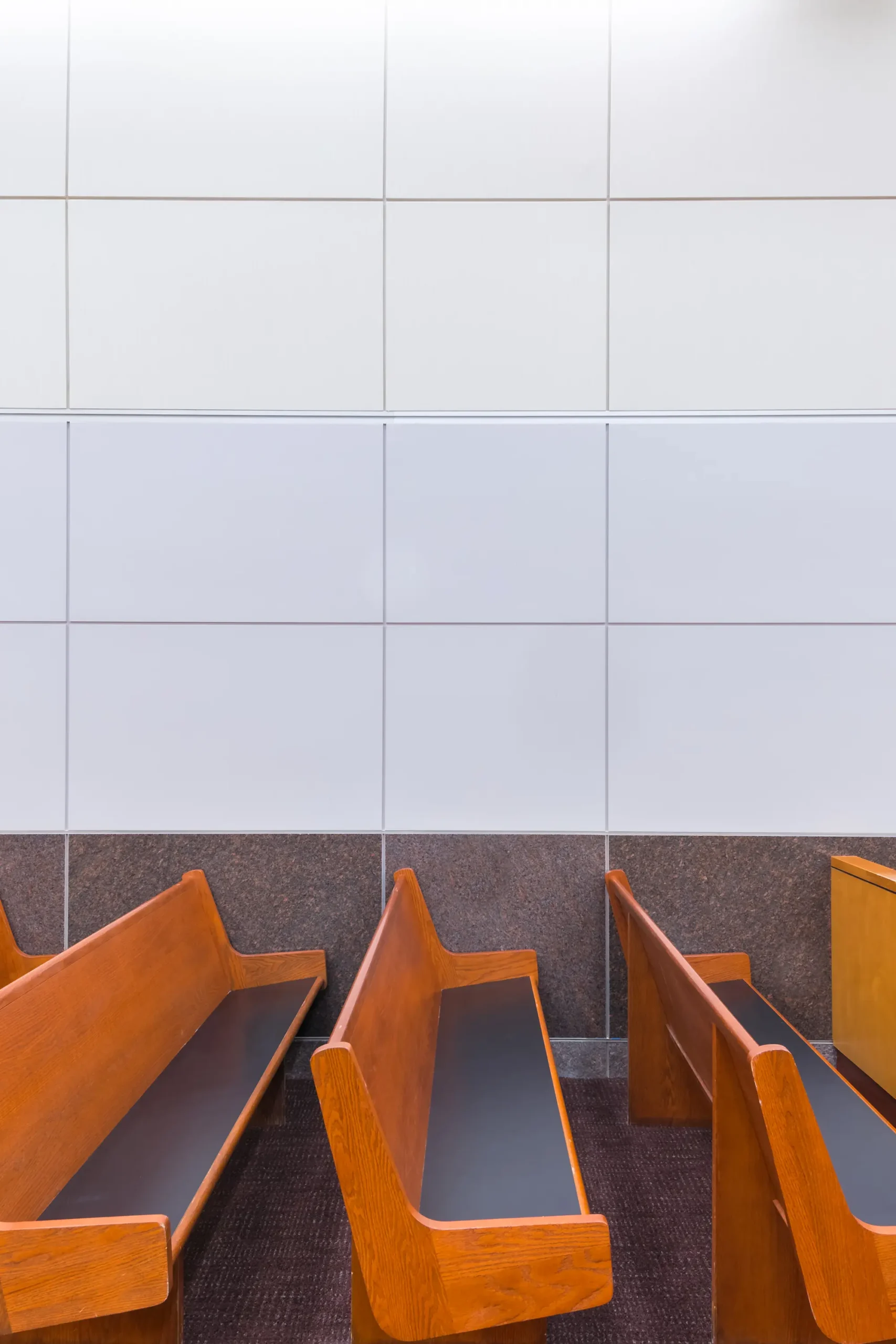 Los bancos de madera se encuentran sobre una alfombra marrón en una sala con paredes alicatadas en blanco y marrón, lo que transmite una atmósfera formal y serena.