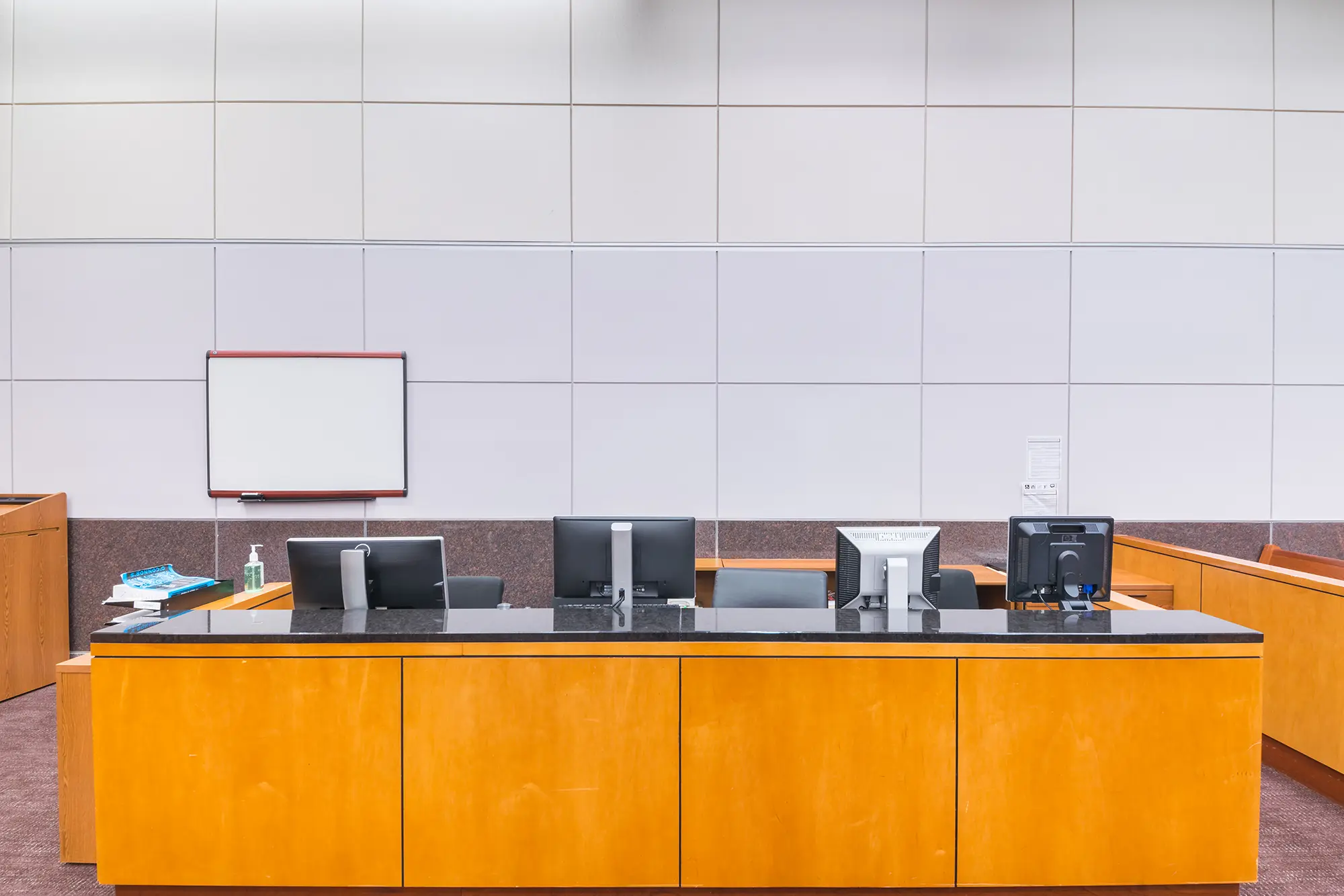 Interior moderno de una sala de audiencias con un estrado de madera para el juez, cuatro monitores de ordenador y una pizarra blanca en la pared, que transmite un ambiente formal y organizado.