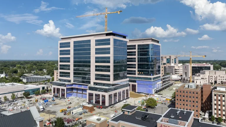 Aerial view of a construction site featuring two modern, glass-fronted office buildings with cranes above. Surrounding structures and clear blue sky.