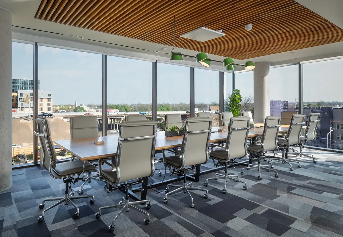 Modern conference room with a long wooden table, twelve sleek chairs, large windows with city views, and ceiling lights, conveying a professional atmosphere.