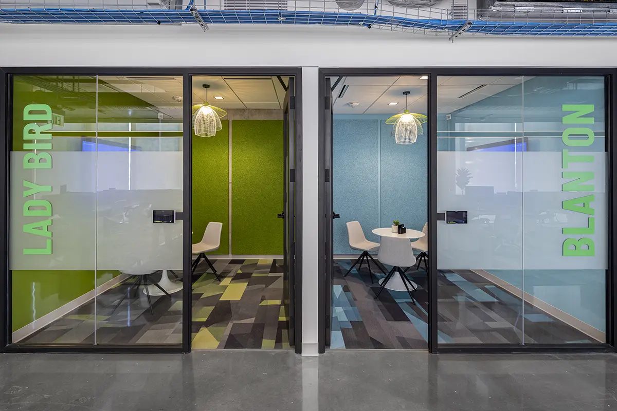 Modern office with two glass meeting rooms labeled "LADY BIRD" and "BLANTON." Rooms have colorful walls, pendant lights, and minimalist furniture.