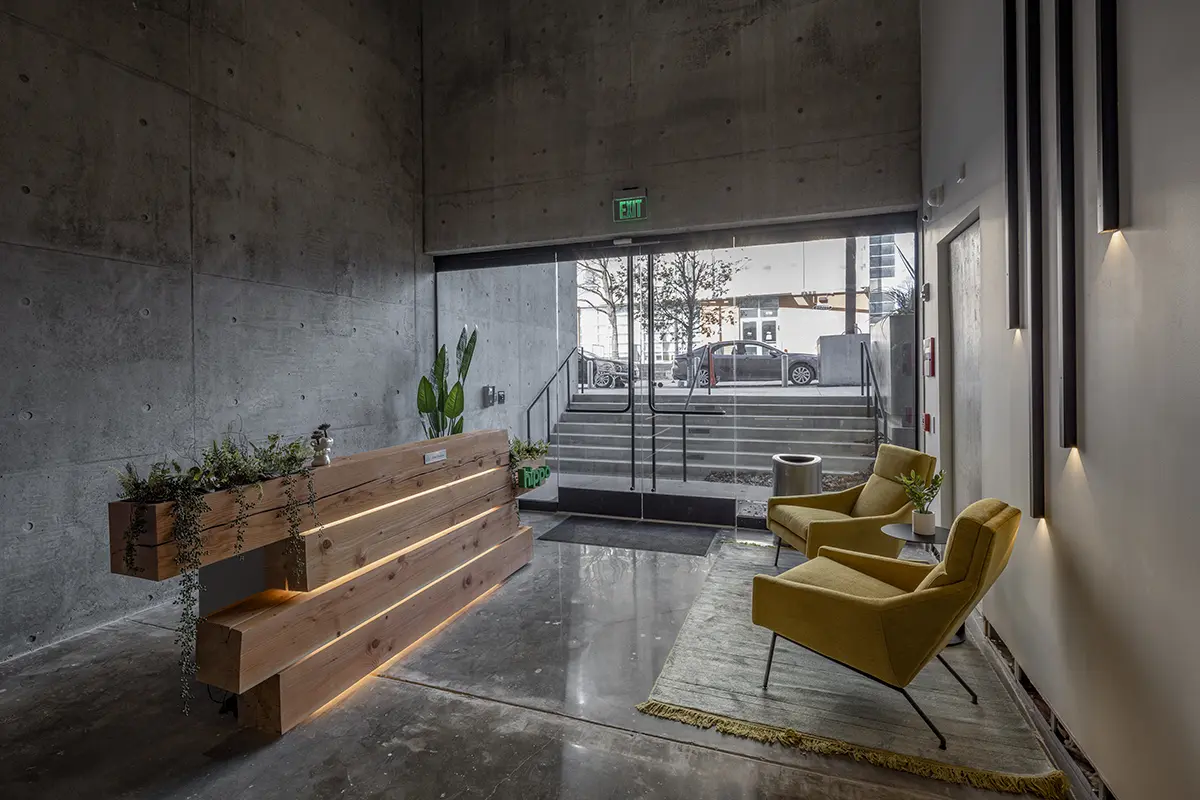 Modern lobby with a sleek wooden reception desk adorned with greenery. Two mustard yellow chairs sit on a rug near a glass door. Minimalist and calm.