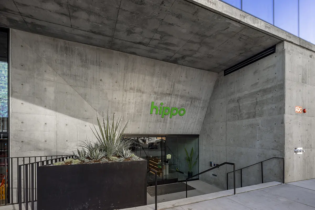 Concrete building entrance with modern design and prominent green "hippo" logo. There is a planter with desert plants next to a glass doorway.