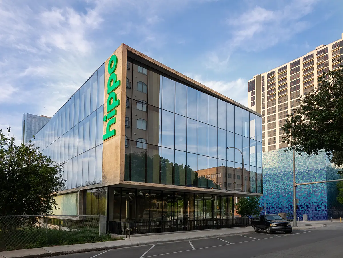 Modern glass building with green "hippo" sign, reflecting a blue sky and urban surroundings. Nearby is a mosaic-tiled wall and a parked black car.
